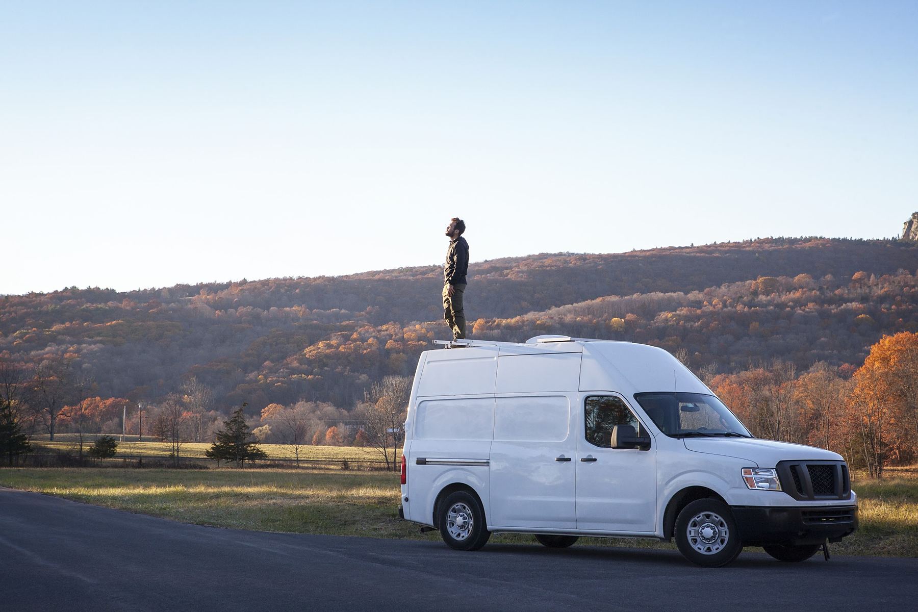 Greg standing on the roof of his first van, a white Nissan NV, in front of autumn hills